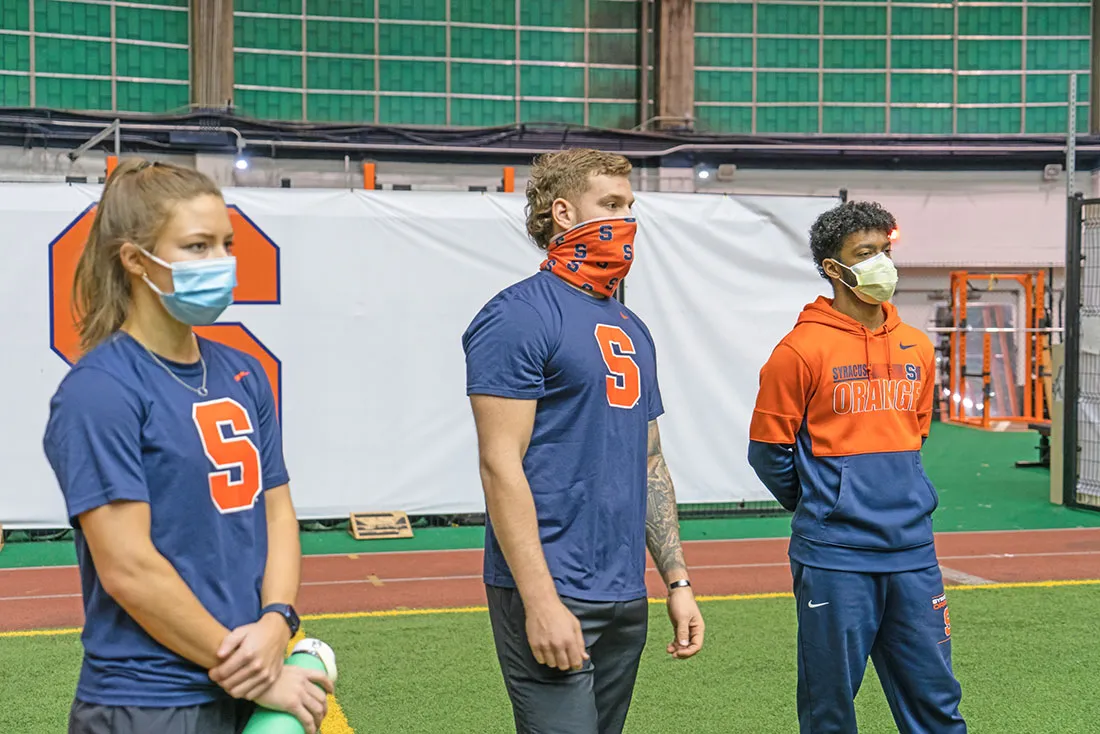 three students dressed in SU shirts standing on athletic field wearing masks