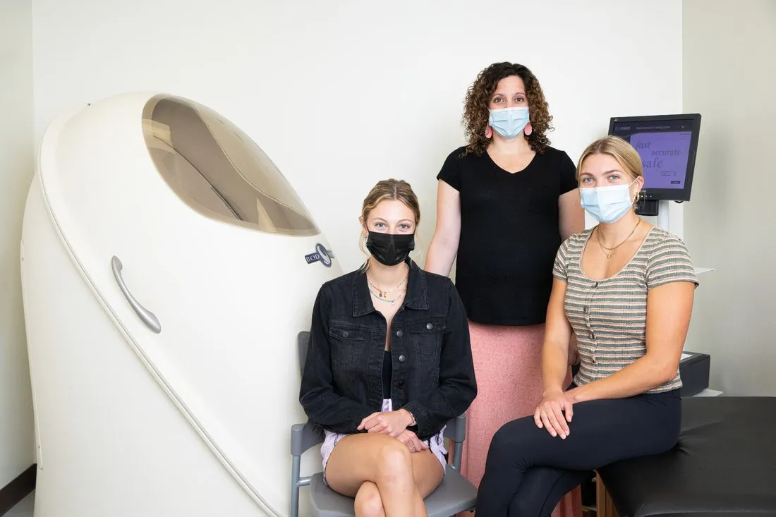 three females wearing masks sitting outside a Bod Pod which analyzed body composition