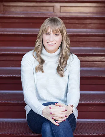 headshot of young woman with long blonde hair and white sweater, sitting on brown stairs