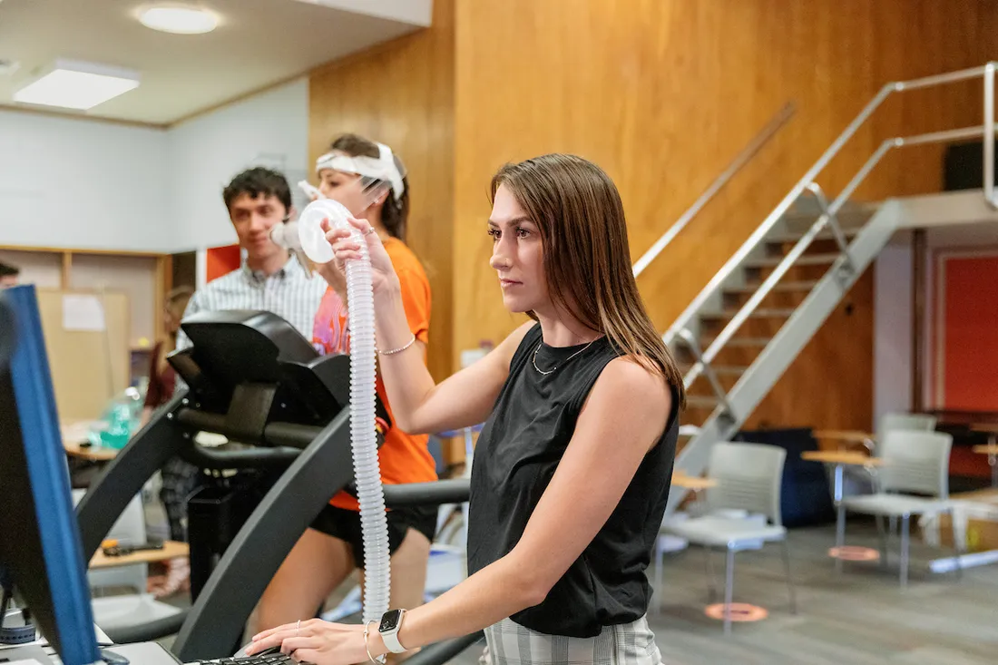 Two students observe a third on a treadmill wearing a metabolic mask