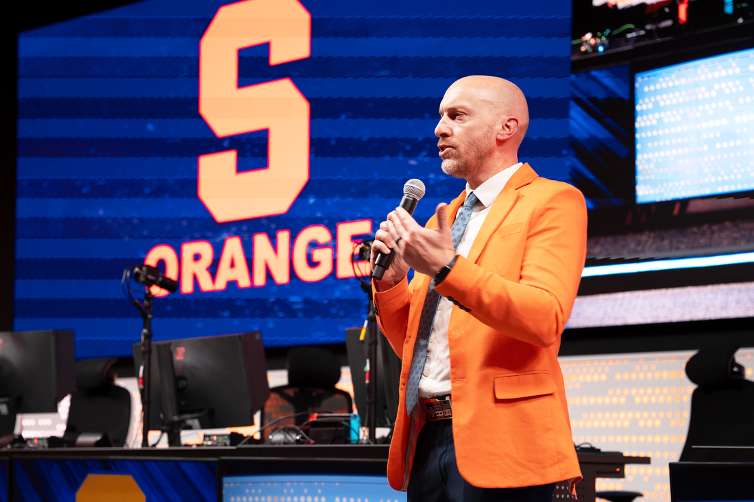 A man in an orange suit speaks into a microphone during a presentation or event.
