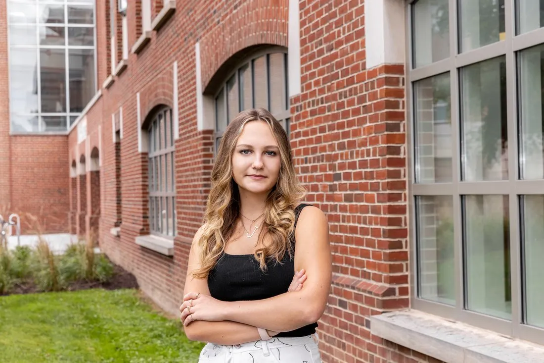 female student with long blonde hair wearing dark top and white pants stands outside brick building