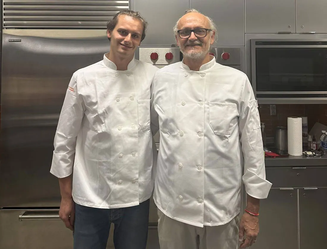 Older man and his adult son standing side by side dressed in chef's white jackets in front of kitchen set up
