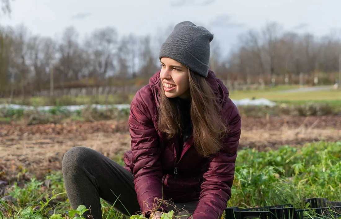 student with long brown hair wearing a brown hat and scarf is bent down in a radish patch