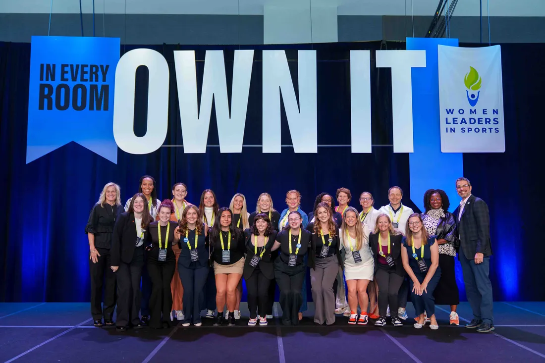 Students, faculty and staff pose with a sign that says "OWN IT" at the Women Leaders in Sports Convention