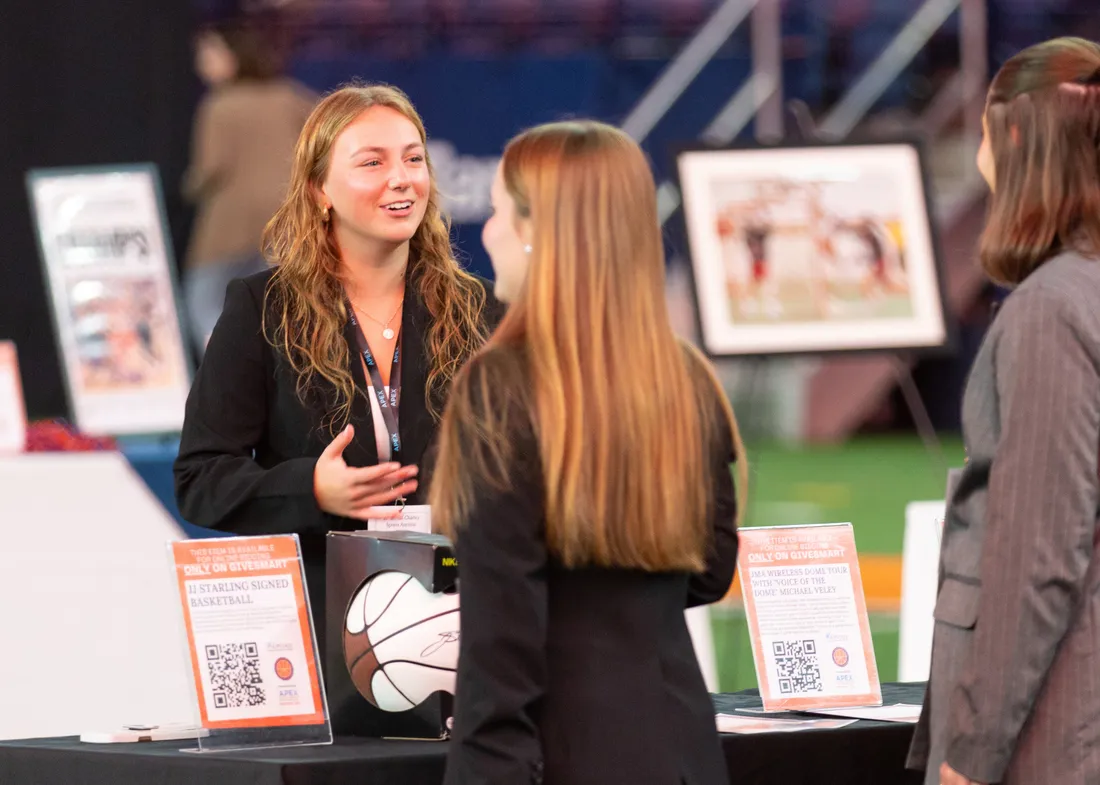 Three women in business attire stand and talk at an indoor event booth with informational signs, a basketball, and framed photos on display.