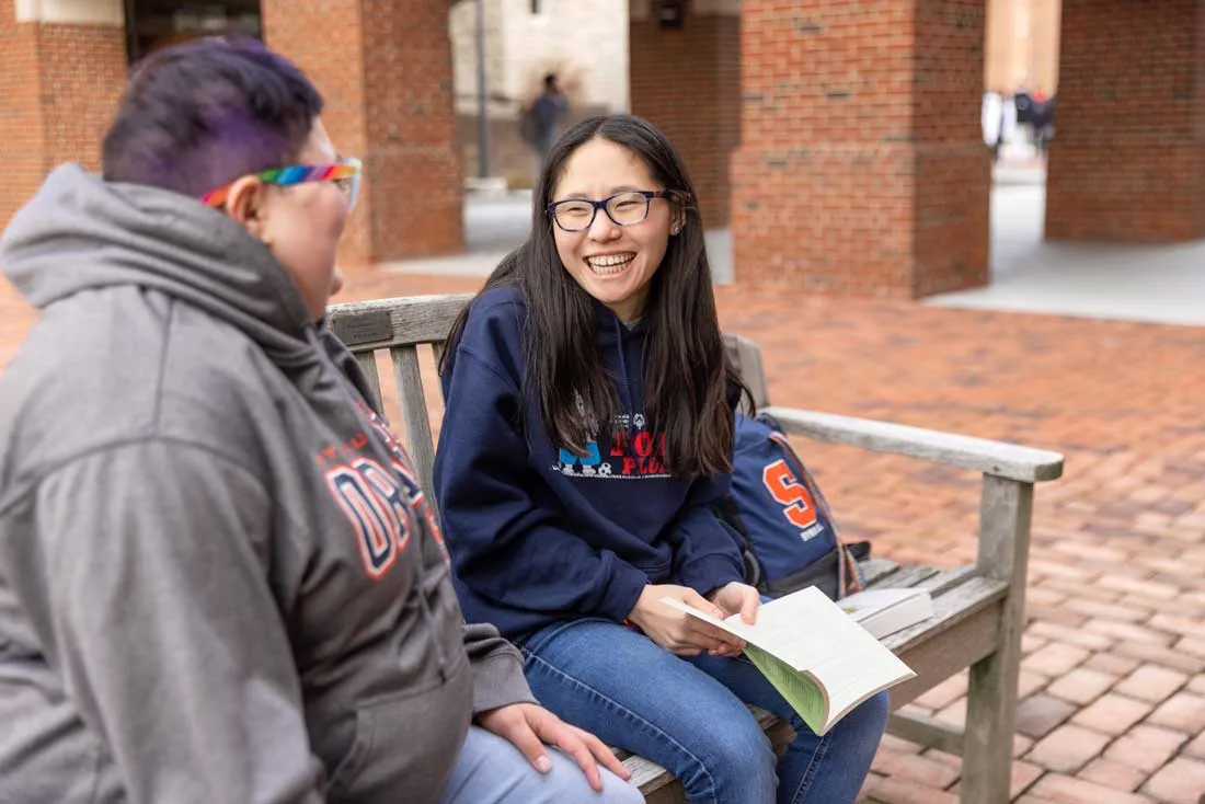 a male and female student share a laugh while sitting on a bench