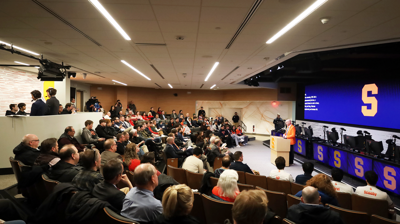 A crowd sits in an auditorium watching a speaker at a podium next to a large screen displaying a yellow "S" on a blue background.