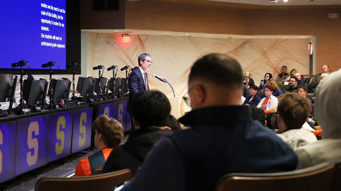 A man delivers a speech to an audience, with a screen behind him showing relevant content.