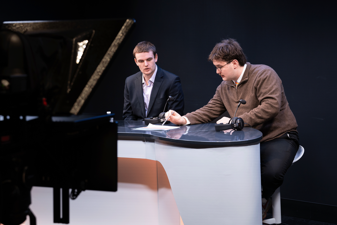 Two men seated at a desk, facing a camera, engaged in conversation or discussion.