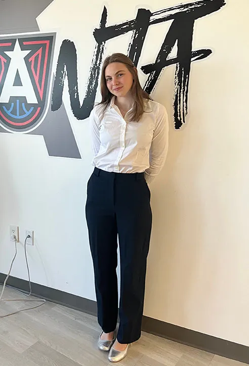 female with long brown hair standing against a wall is wearing dark pants and light colored shirt