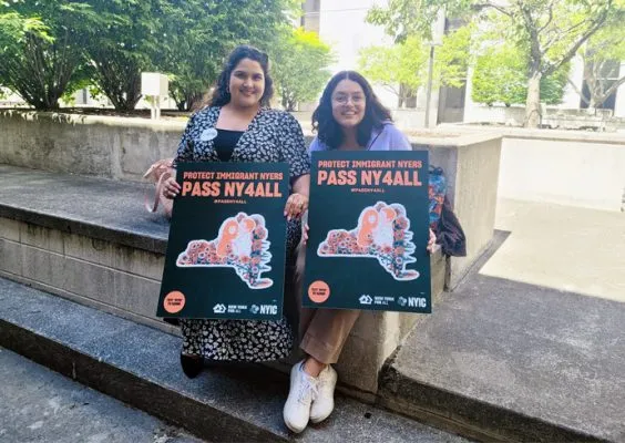 two students sitting on concrete wall holding signs that say Pass NY4All