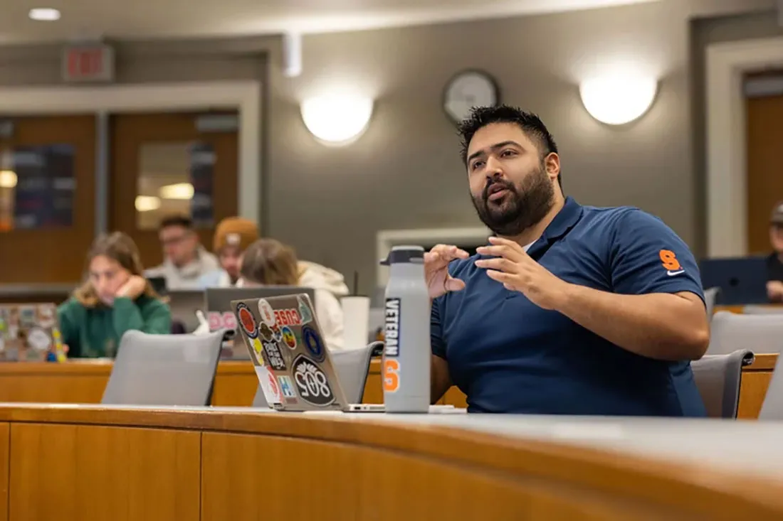 bearded man sitting at table speaking to a class