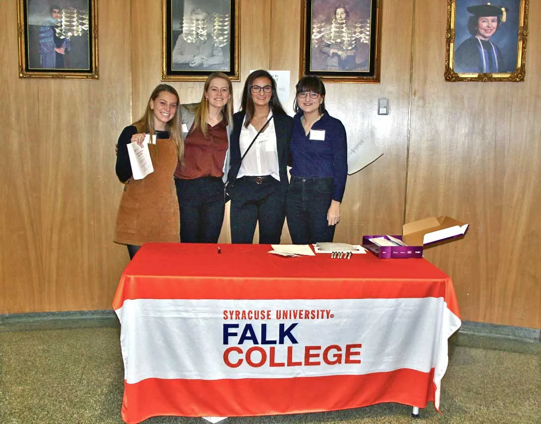 four young woman standing in front of a table with Falk College banner