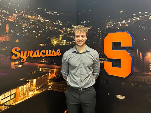 student tyler standing in front of Syracuse University logo