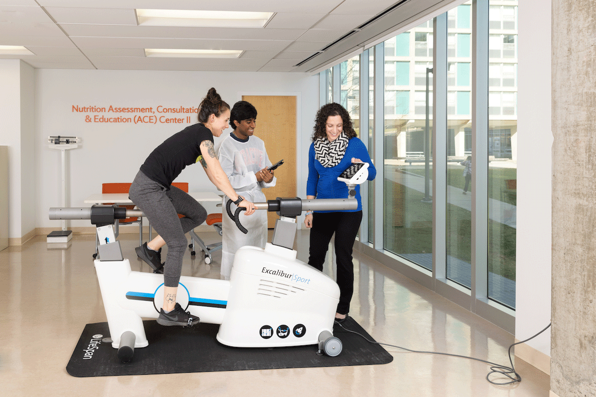 student riding exercise bike with attendant and faculty member observing
