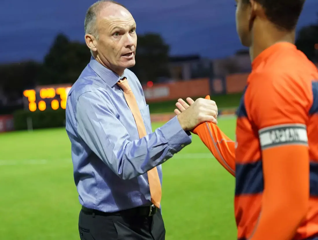 mens soccer coach in blue shirt and tie shakes hands with captain wearing an orange and blue shirt