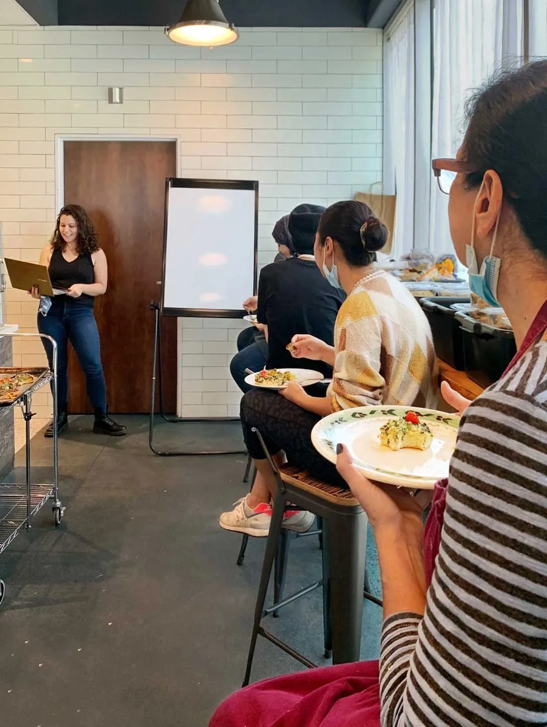 four immigrant women sit at a counter eating while a young woman off to the left is looking at her computer screen