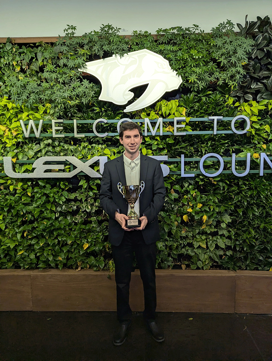 Austin Ambler holding a trophy standing in front of welcome sign covered wih greenery