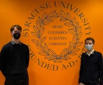 two students standing by the SU logo on an orange wall