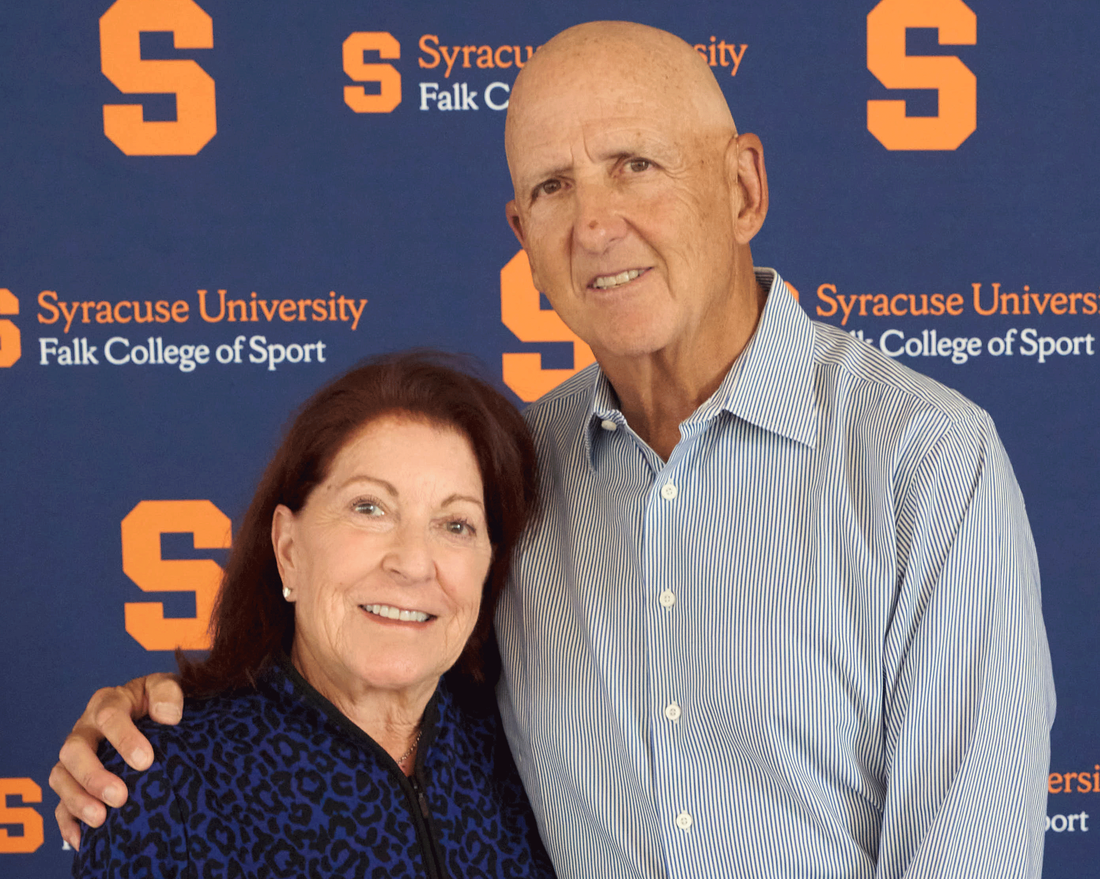 David Falk and Rhonda Falk standing side by side in front of blue an orange wall with Falk and SU logos
