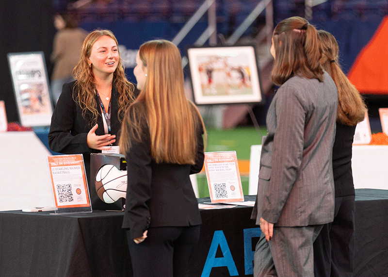Three women in business attire stand and talk at an indoor event booth with informational signs, a basketball, and framed photos on display.