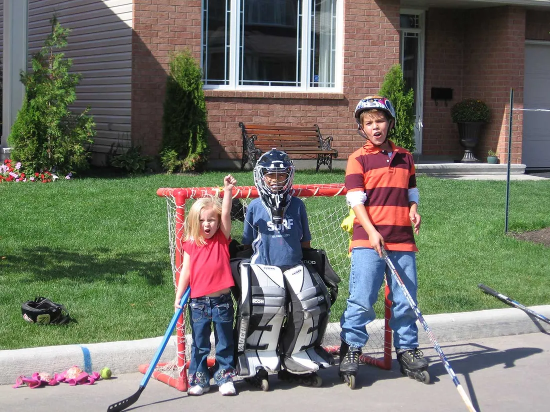 three young kids stand in front of hockey net. The middle one wearing goalie gear